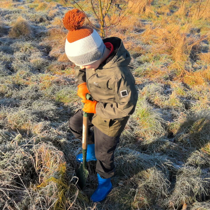 Little boy planting a tree in orange bobble hat and blue wellies