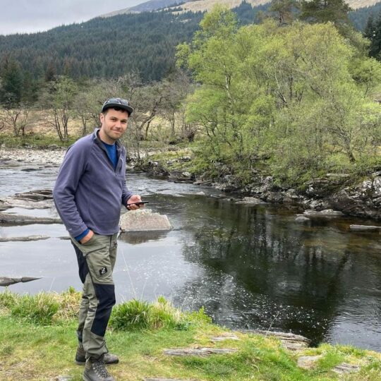 Man smiling, standing in front of lake with trees in the hills
